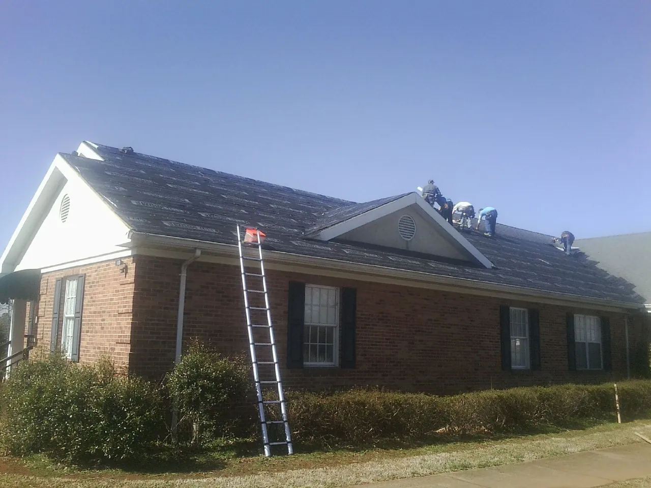 Crew tearing shingles off a brick ranch roof with a ladder set — Metal Roof Repair work by our crew in West Kingston